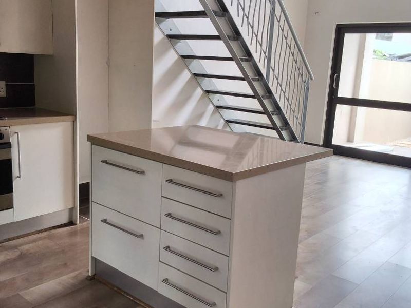 Open-plan living area featuring a modern kitchen island, laminate flooring, exposed timber ceiling beams, and a metal staircase leading to an upper level.