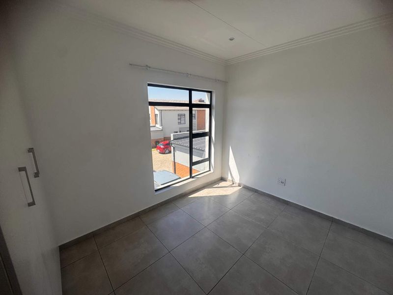 A bright, empty bedroom featuring neutral tiled flooring, built-in cupboards, and a large window providing natural light and views of the complex.