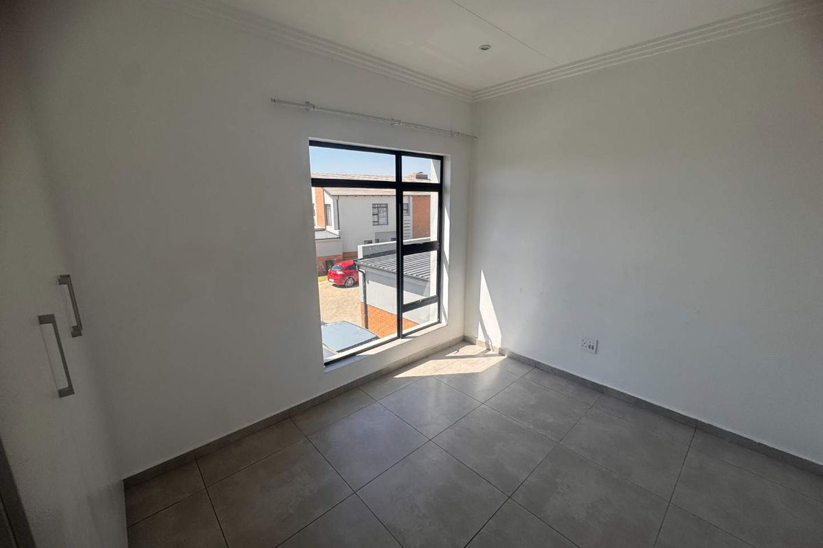 A bright, empty bedroom featuring neutral tiled flooring, built-in cupboards, and a large window providing natural light and views of the complex.