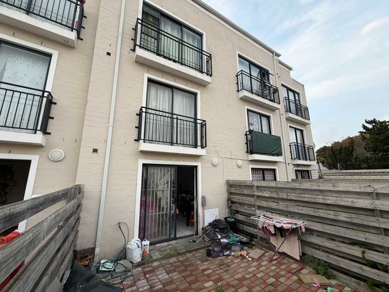 Rear view of a multi-story apartment building featuring brick facade, private balconies, and a paved ground-level patio area enclosed by timber fencing.