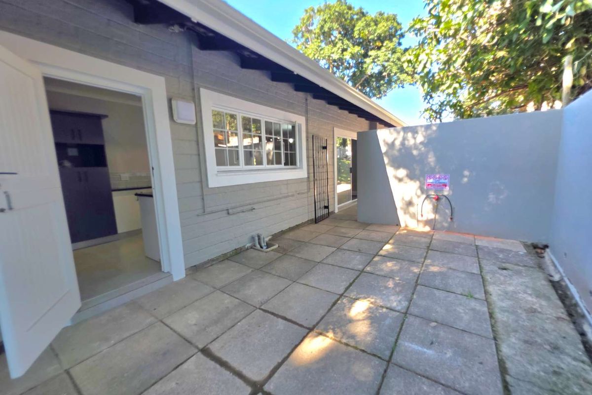 A paved rear courtyard area featuring a grey exterior facade, white window frames, and direct access to the kitchen and living spaces.