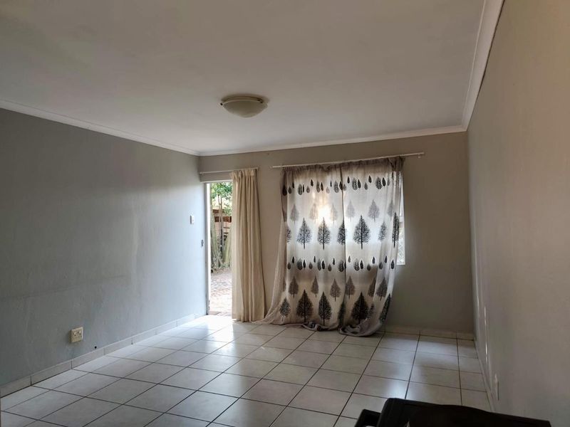 A bright, empty living room featuring neutral tiled flooring, a ceiling light fixture, and direct access to an outdoor area through a glass door.