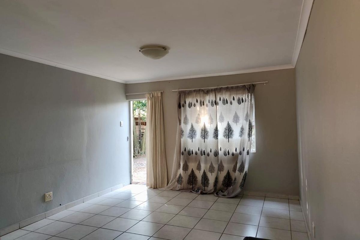 A bright, empty living room featuring neutral tiled flooring, a ceiling light fixture, and direct access to an outdoor area through a glass door.