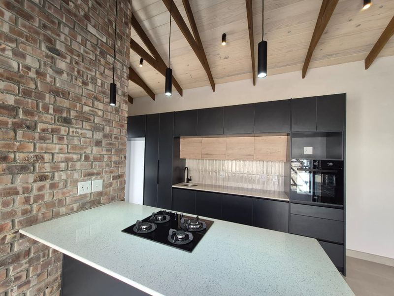 Modern kitchen featuring a striking exposed brick wall, vaulted timber ceiling, sleek dark cabinetry, stone countertops, and integrated gas hob and oven.
