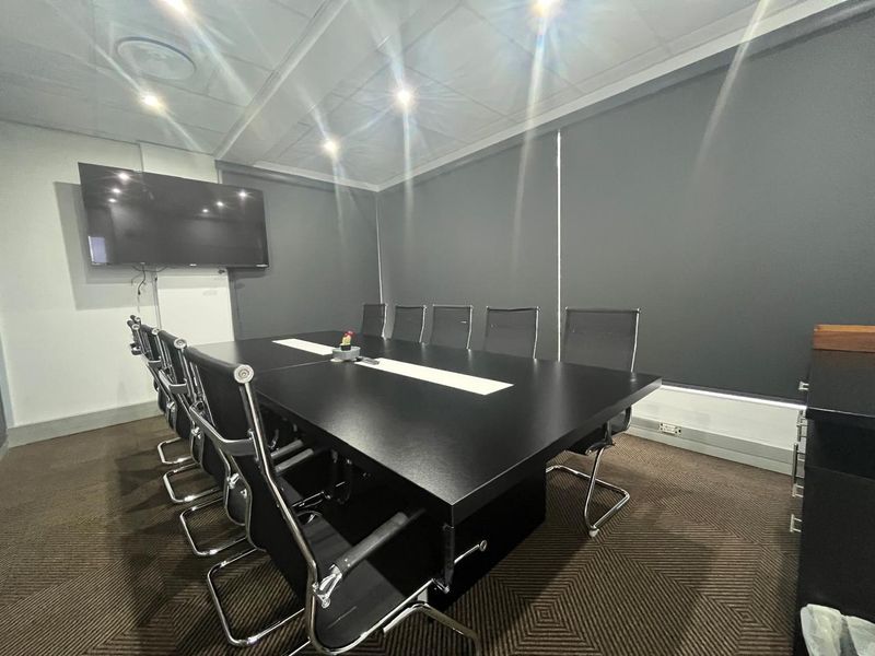 A professional boardroom or meeting room featuring a large conference table, wall-mounted display screen, recessed lighting, and modern roller blinds.