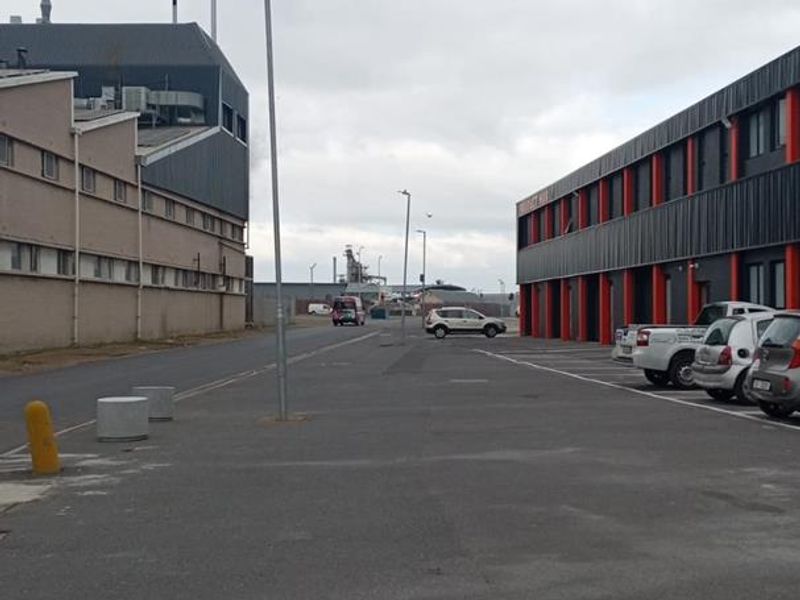 View of an industrial complex access road featuring asphalt paving, street lighting, and adjacent commercial building facades with parking facilities.
