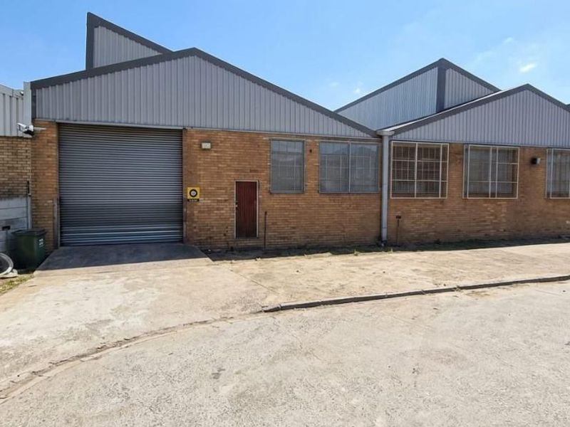 Front exterior view of an industrial warehouse unit featuring a roller shutter door, brick facade, and metal roofing with a concrete driveway.