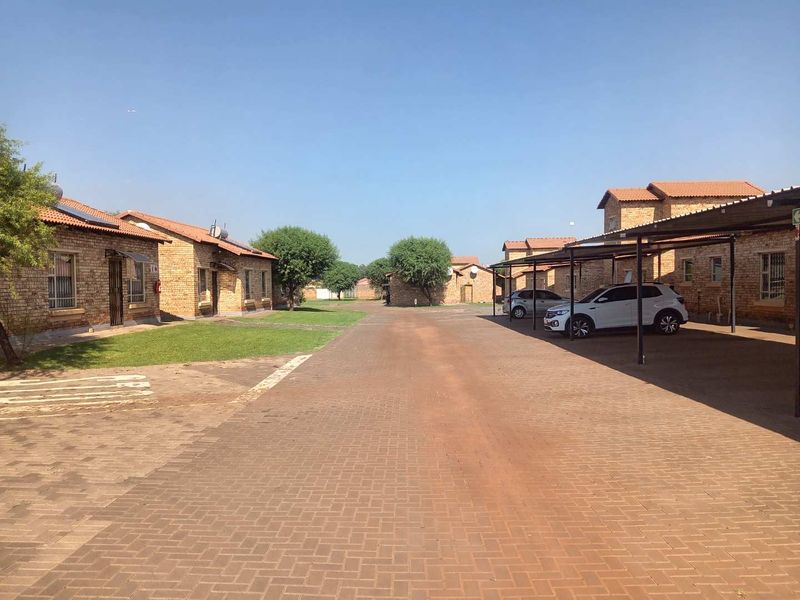 A view of the townhouse complex street, featuring paved driveways, covered carports, and brick-faced residential units with solar geysers.