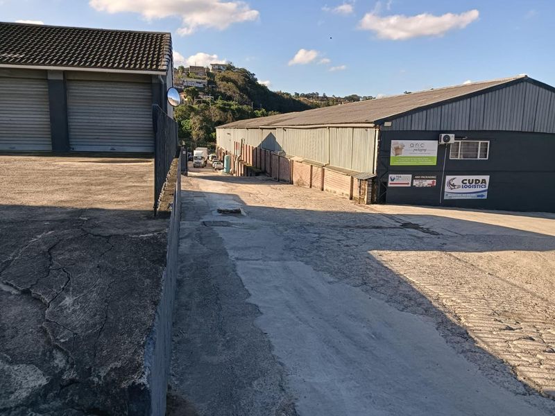 Exterior view of an industrial property featuring a concrete driveway, metal roller shutter doors, and corrugated metal roofing on the warehouse structure.