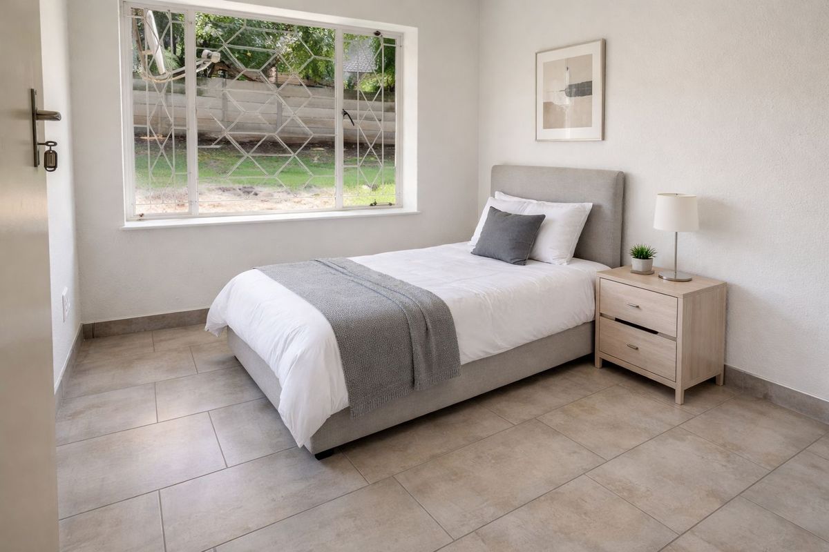 A bright bedroom featuring neutral tiled flooring and a large window with security bars, offering a clean and functional living space.