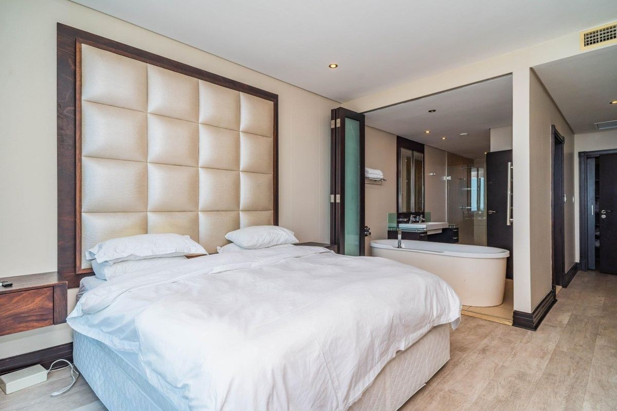 Modern bedroom featuring an integrated open-plan bathroom with a freestanding bathtub, vanity, and glass-enclosed shower, complemented by a large upholstered headboard.