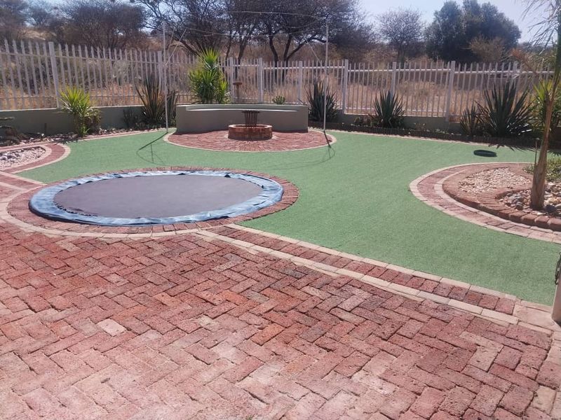 A well-maintained landscaped garden featuring brick paving, artificial turf, a built-in fire pit with a curved concrete bench, and secure perimeter fencing.