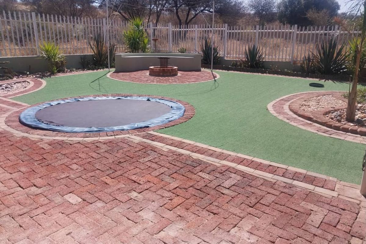 A well-maintained landscaped garden featuring brick paving, artificial turf, a built-in fire pit with a curved concrete bench, and secure perimeter fencing.