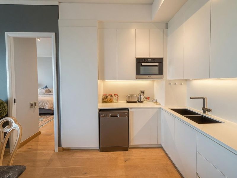 Modern kitchen featuring sleek white cabinetry, quartz countertops, integrated microwave, and high-quality hardwood flooring, with a seamless transition to the adjacent bedroom.