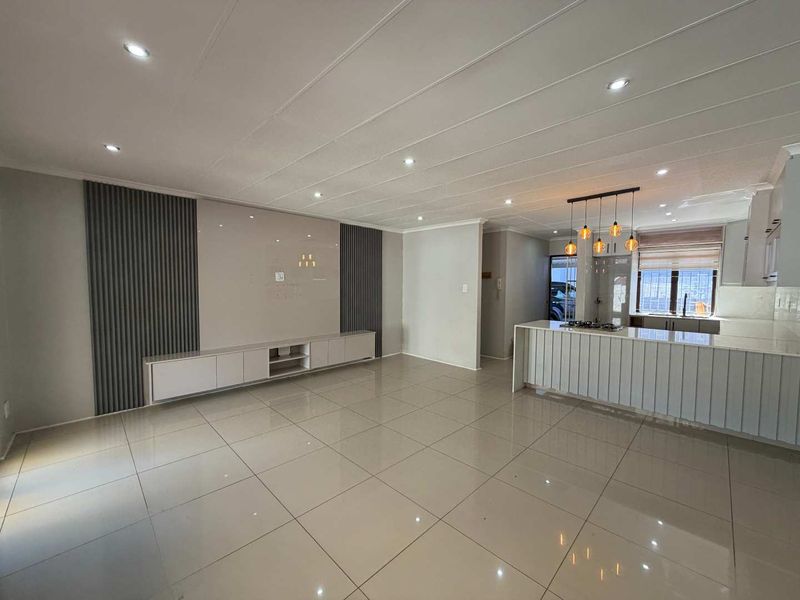 Spacious open-plan living area featuring modern tiled floors, recessed lighting, a sleek built-in media unit with decorative wall paneling, and a functional kitchen island.