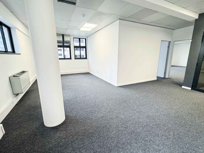 A bright, open-plan office space featuring neutral carpeting, a suspended ceiling with recessed lighting, and a wall-mounted HVAC unit.