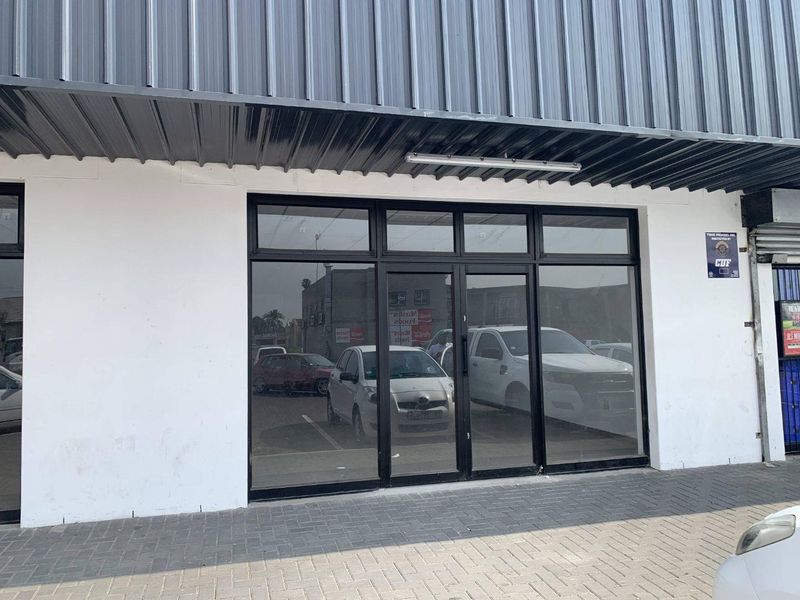 Front facade of a retail unit featuring a black aluminum-framed glass storefront, a metal corrugated awning, and a paved pedestrian walkway.