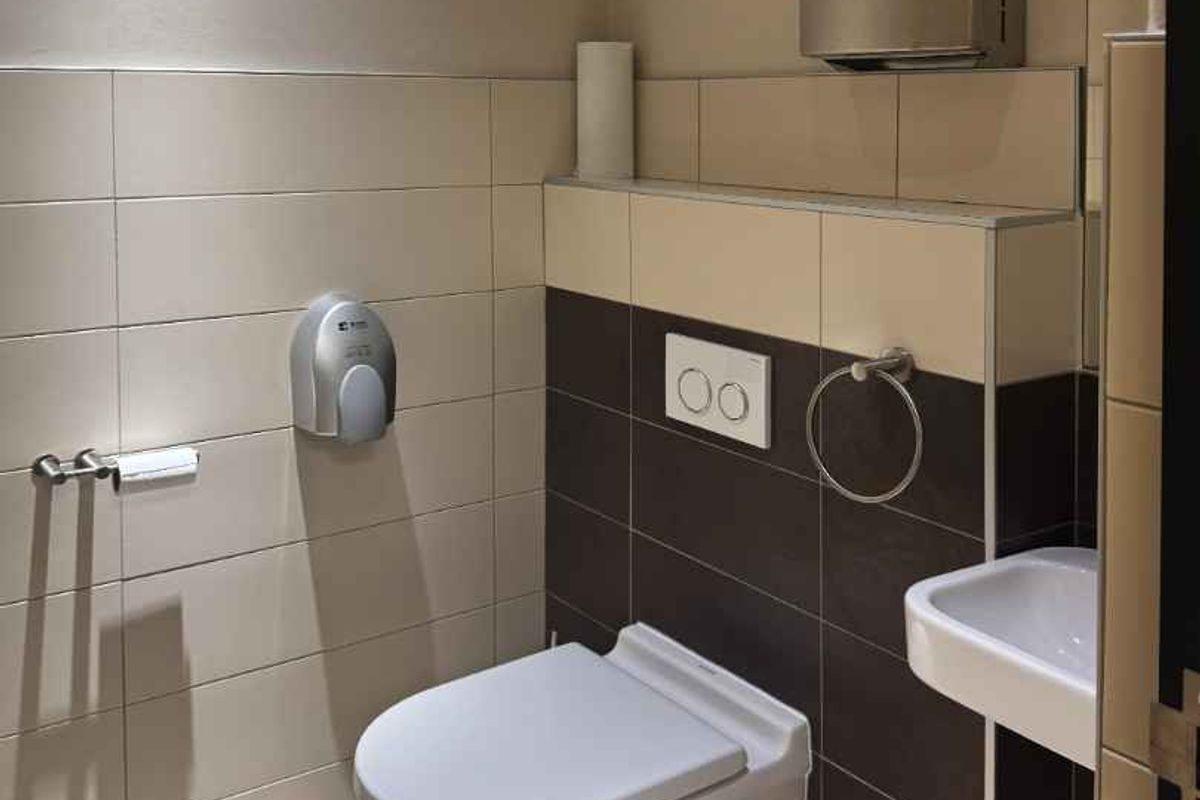A modern, clean office restroom featuring a wall-mounted toilet, small basin, and integrated paper towel dispenser with neutral-toned tiled walls.