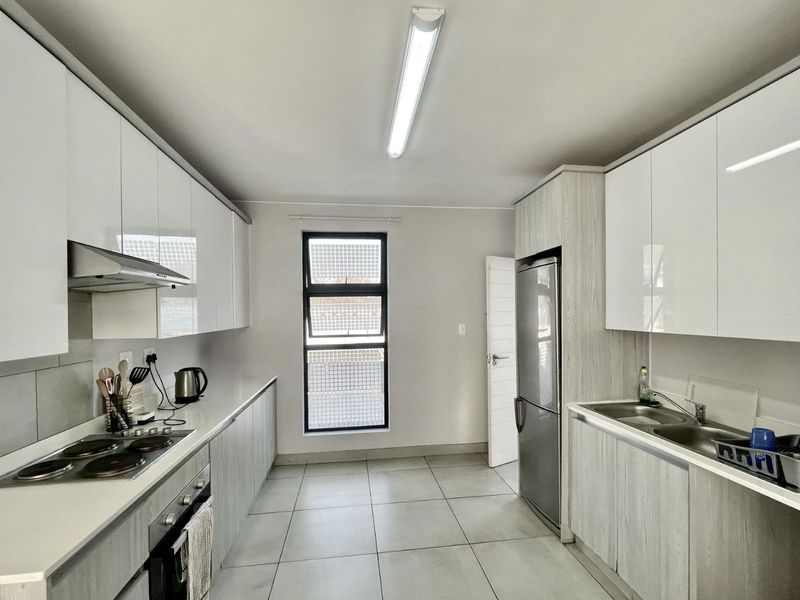 Modern galley-style kitchen featuring white high-gloss cabinetry, integrated electric hob, oven, extractor fan, and double sink with tiled flooring.