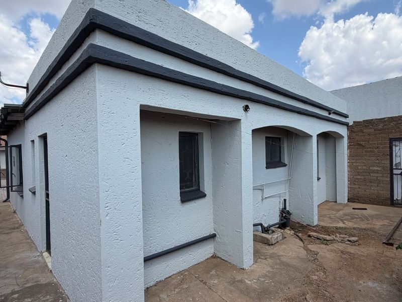 Rear exterior view of the property featuring a white rendered facade, security gate, and paved outdoor area with exposed external plumbing.