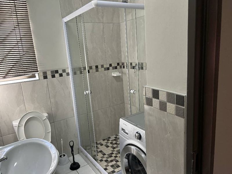 A functional bathroom featuring a glass-enclosed shower, pedestal basin, toilet, and neutral wall tiling with a decorative mosaic border.