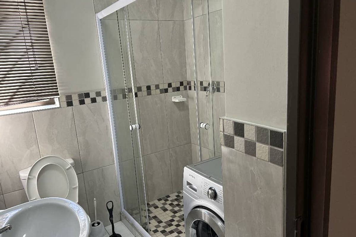 A functional bathroom featuring a glass-enclosed shower, pedestal basin, toilet, and neutral wall tiling with a decorative mosaic border.