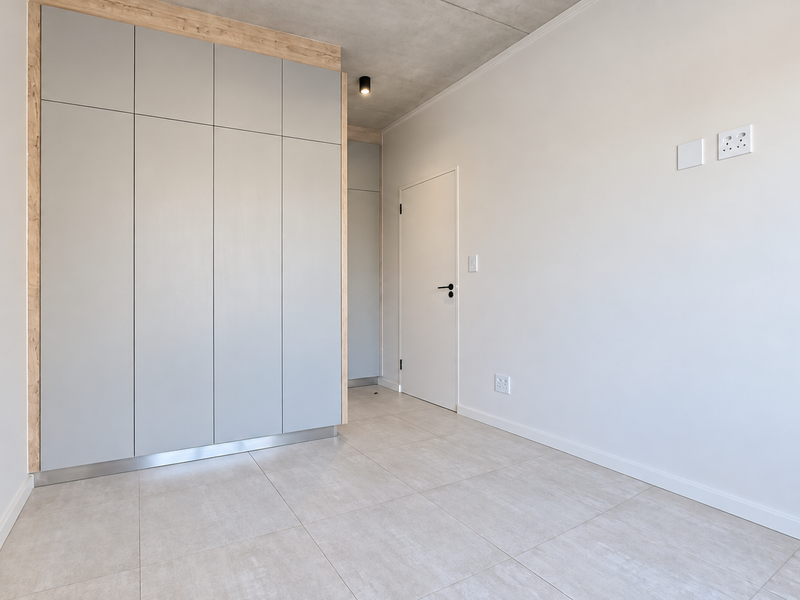 A modern, empty bedroom featuring sleek built-in cupboards, neutral tiled flooring, and an exposed concrete ceiling, offering a clean and contemporary living space.