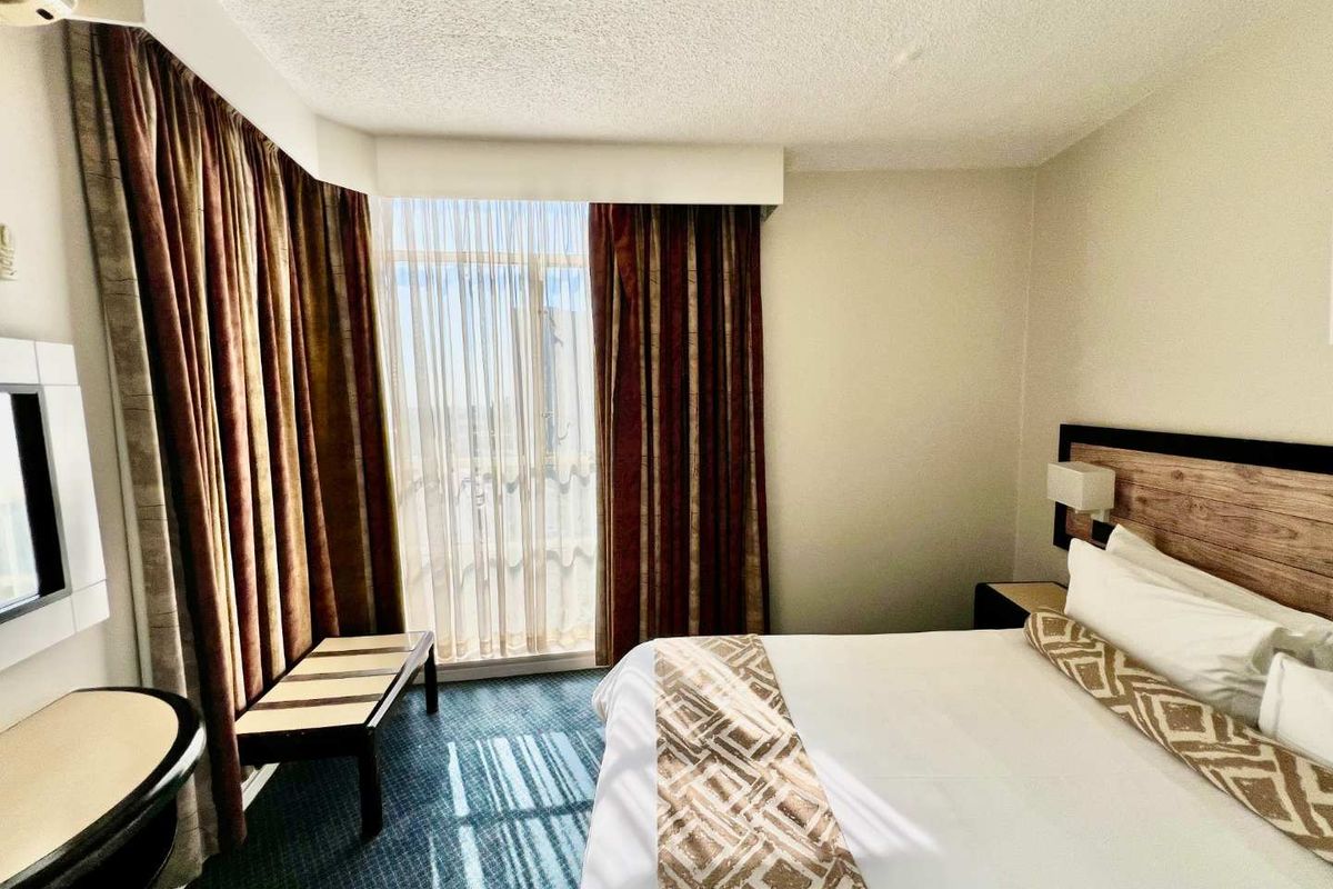 A bedroom featuring a large window with floor-to-ceiling curtains, wall-mounted headboard, and carpeted flooring.