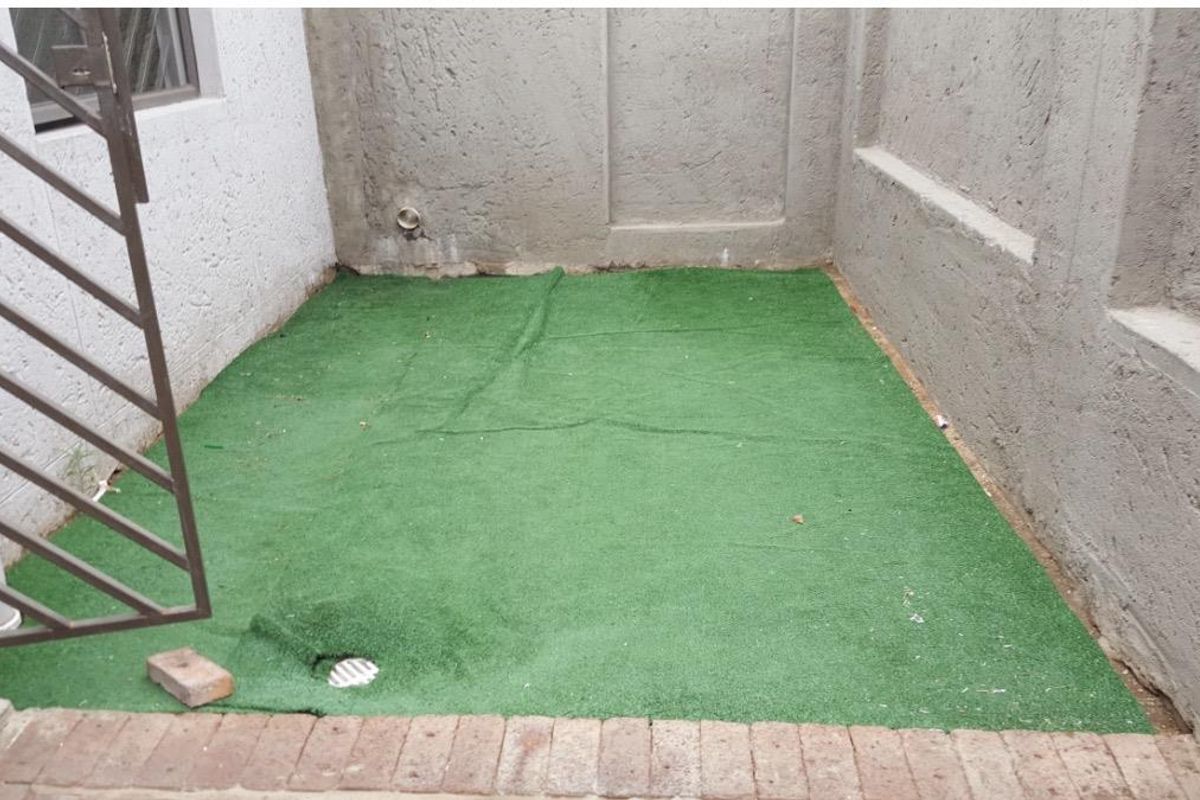 A small, enclosed rear courtyard area featuring artificial turf, plastered boundary walls, and a metal security gate.