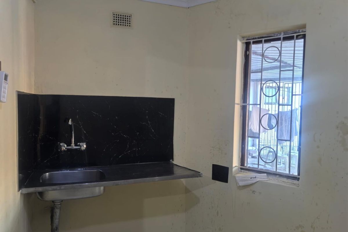 A basic kitchen area featuring a stainless steel sink with a black splashback and security bars on the window.