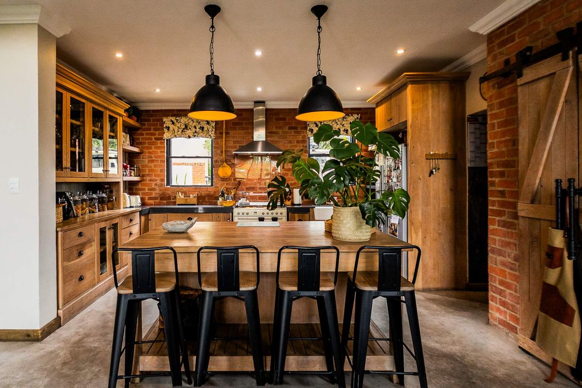 A rustic-style kitchen featuring exposed brick walls, custom wooden cabinetry, a central island, and modern industrial pendant lighting.