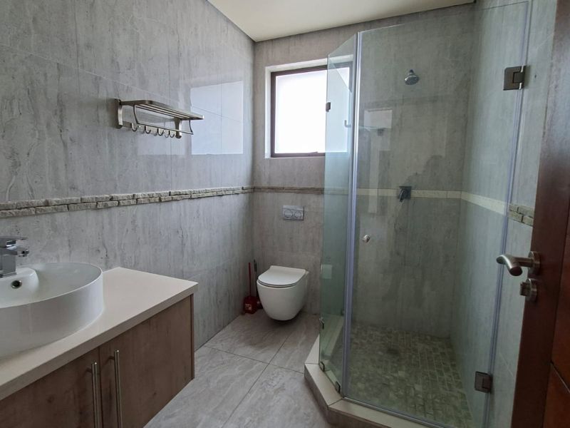 A modern bathroom featuring a vessel sink on a vanity cabinet, a wall-mounted toilet, and a glass-enclosed shower with neutral stone-look tiling.