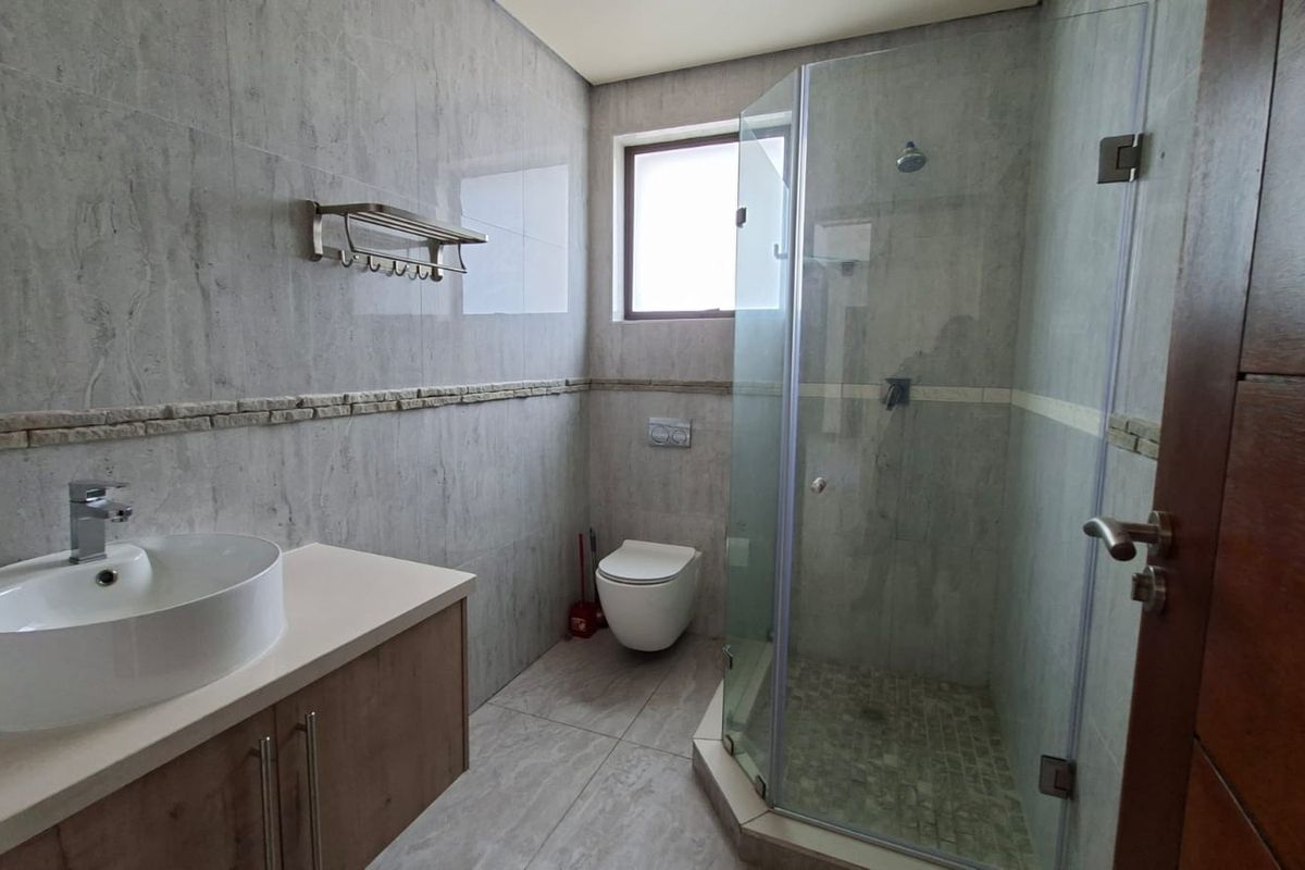 A modern bathroom featuring a vessel sink on a vanity cabinet, a wall-mounted toilet, and a glass-enclosed shower with neutral stone-look tiling.