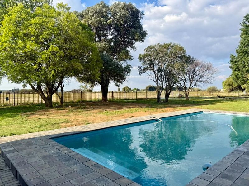 A well-maintained swimming pool with paved surround, set within a spacious garden featuring mature trees and secure perimeter fencing.