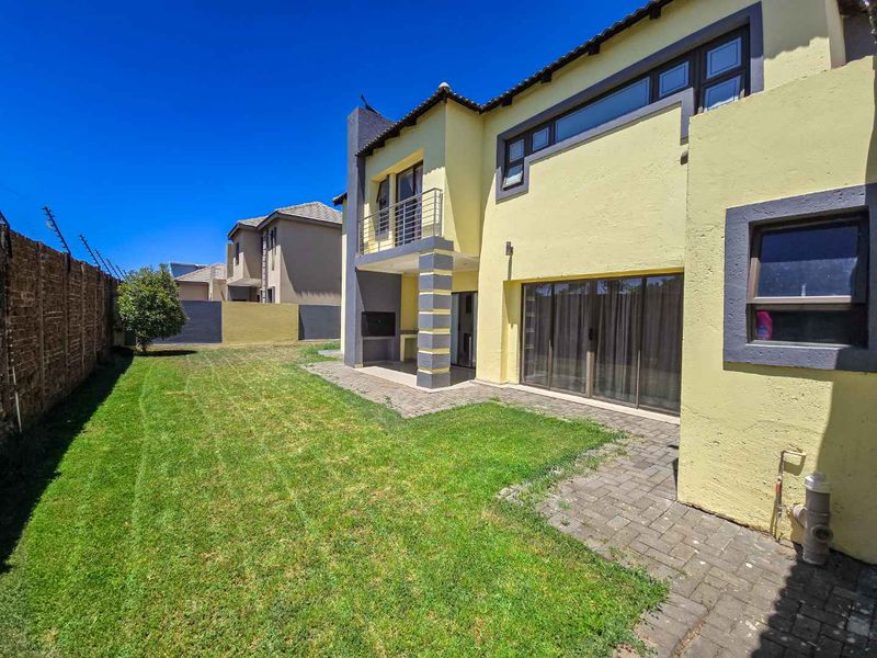Rear view of the townhouse featuring a paved patio, built-in braai, balcony, and a private garden area enclosed by boundary walls.