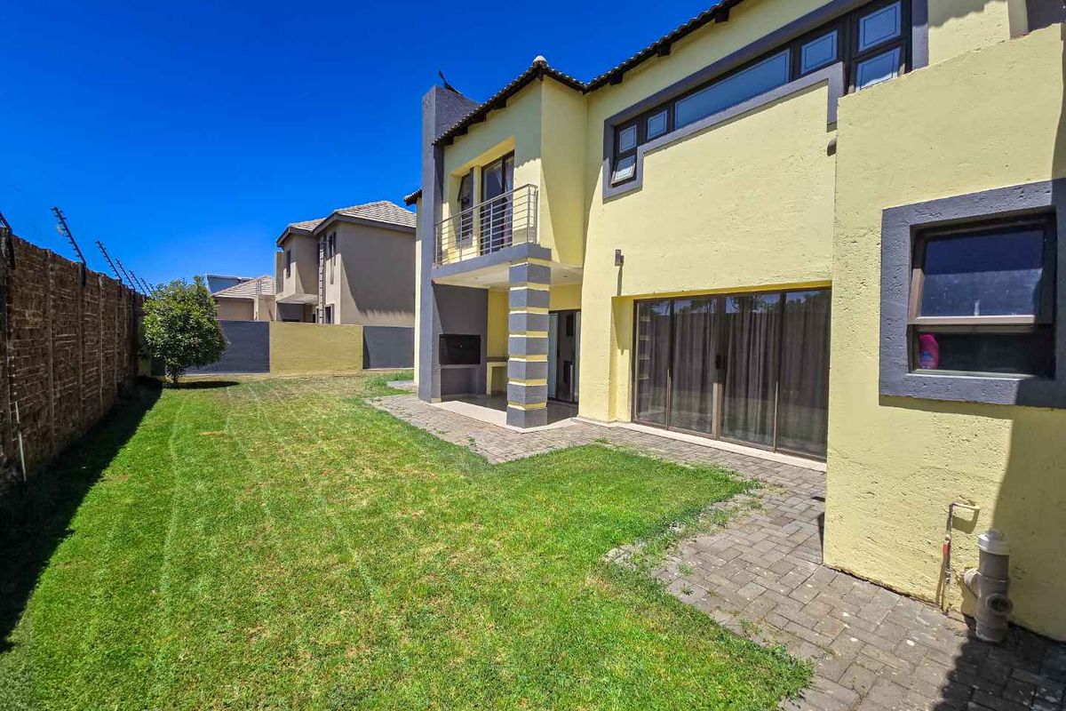 Rear view of the townhouse featuring a paved patio, built-in braai, balcony, and a private garden area enclosed by boundary walls.