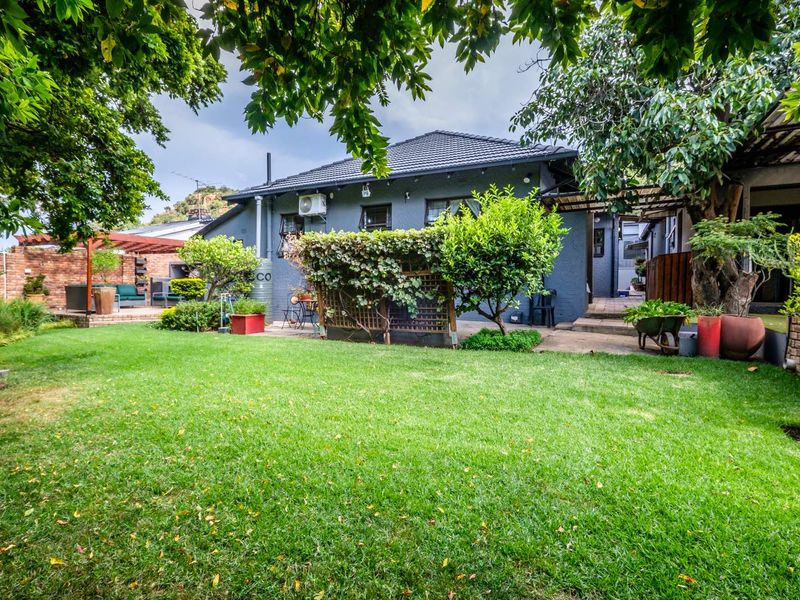 A well-maintained rear garden featuring a lush lawn, mature trees, a paved patio area with a timber pergola, and a secure brick boundary wall.