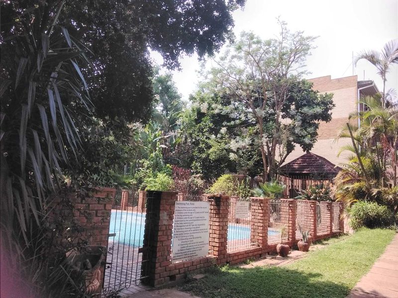 A communal swimming pool area featuring a brick boundary wall, metal security fencing, and established garden surroundings within the apartment complex.