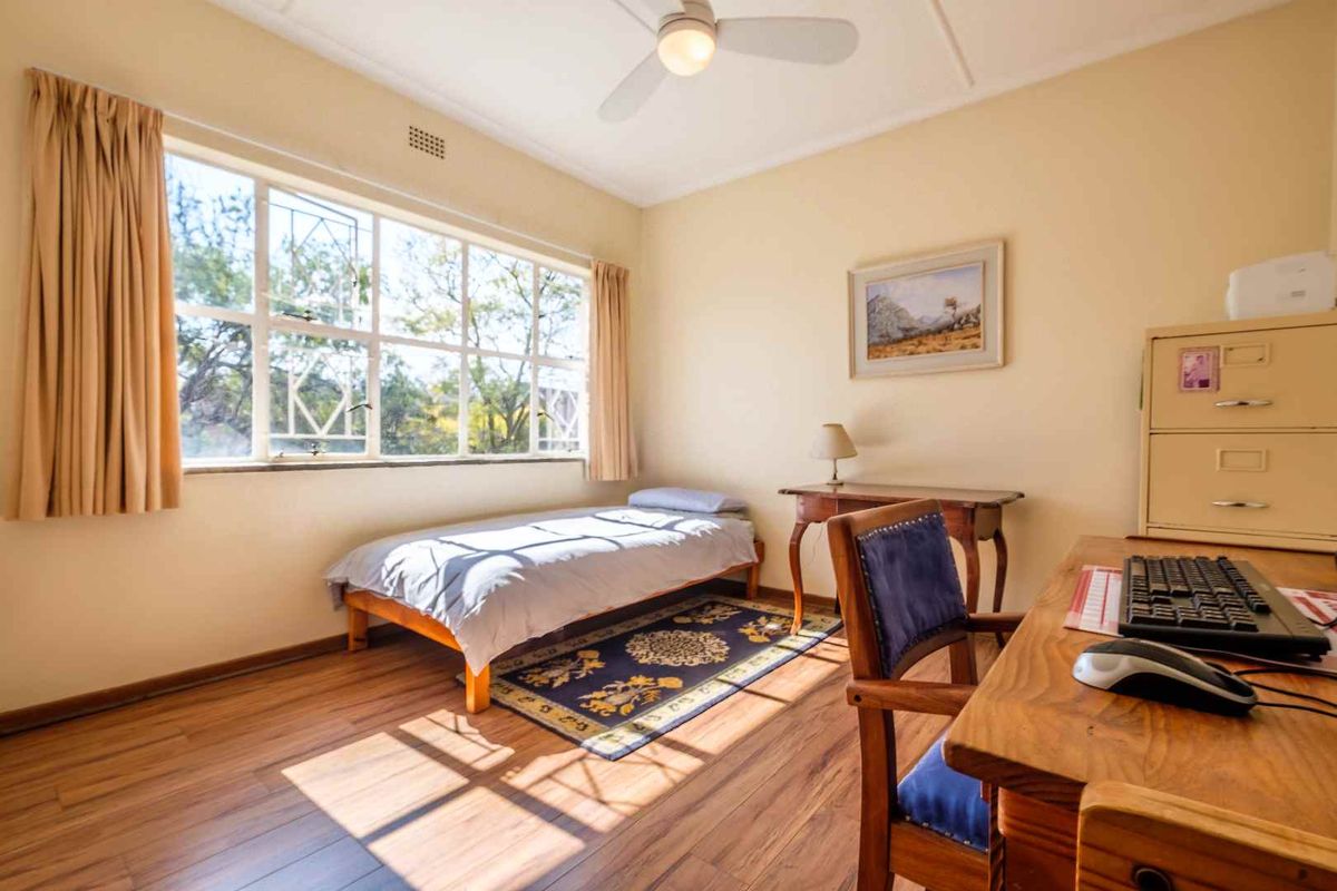 A functional bedroom featuring laminate flooring, a ceiling fan, and a large window with security bars, providing ample natural light.