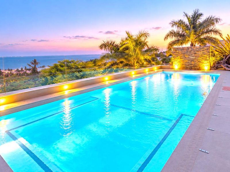 A beautifully illuminated swimming pool area featuring a stone accent wall, glass balustrade, and stunning sunset ocean views.