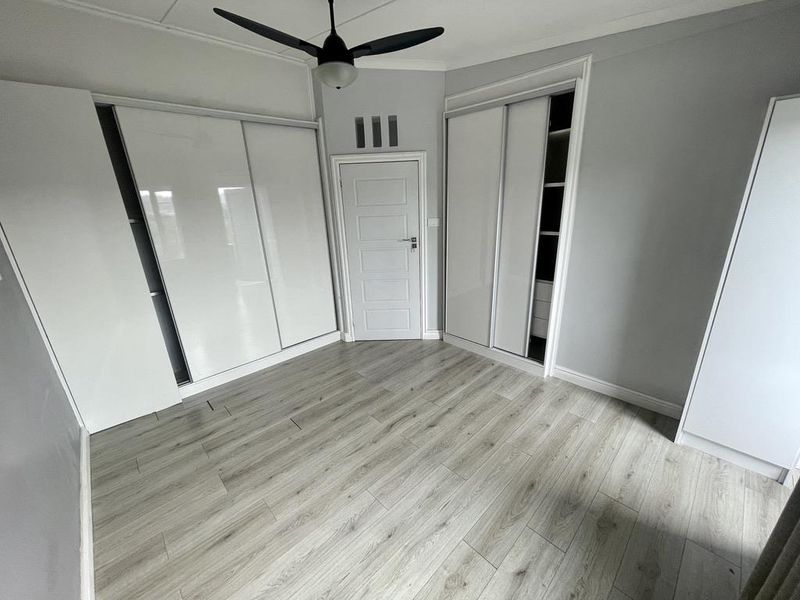 A bright, empty bedroom featuring modern laminate flooring, neutral grey walls, and extensive built-in cupboard storage with a ceiling fan.
