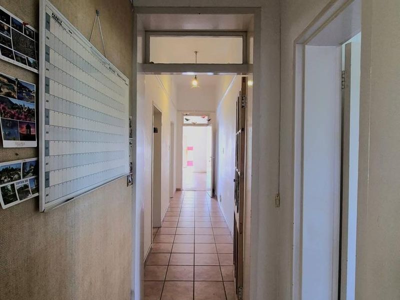 A long, tiled hallway connecting multiple rooms, featuring a transom window above the doorway and dated wall finishes.