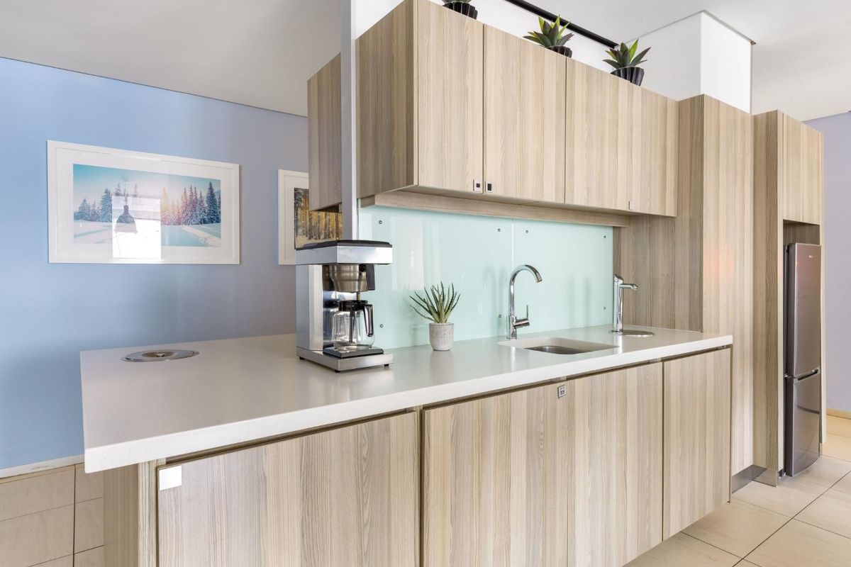 A modern office kitchenette featuring light wood-grain cabinetry, a sleek stone countertop, a glass splashback, and a built-in sink with a chrome faucet.