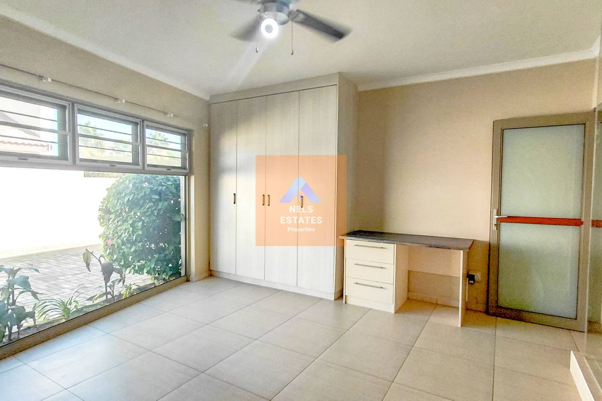 A bright bedroom featuring built-in cupboards, a fitted desk, tiled flooring, and a ceiling fan, with large windows overlooking the exterior.