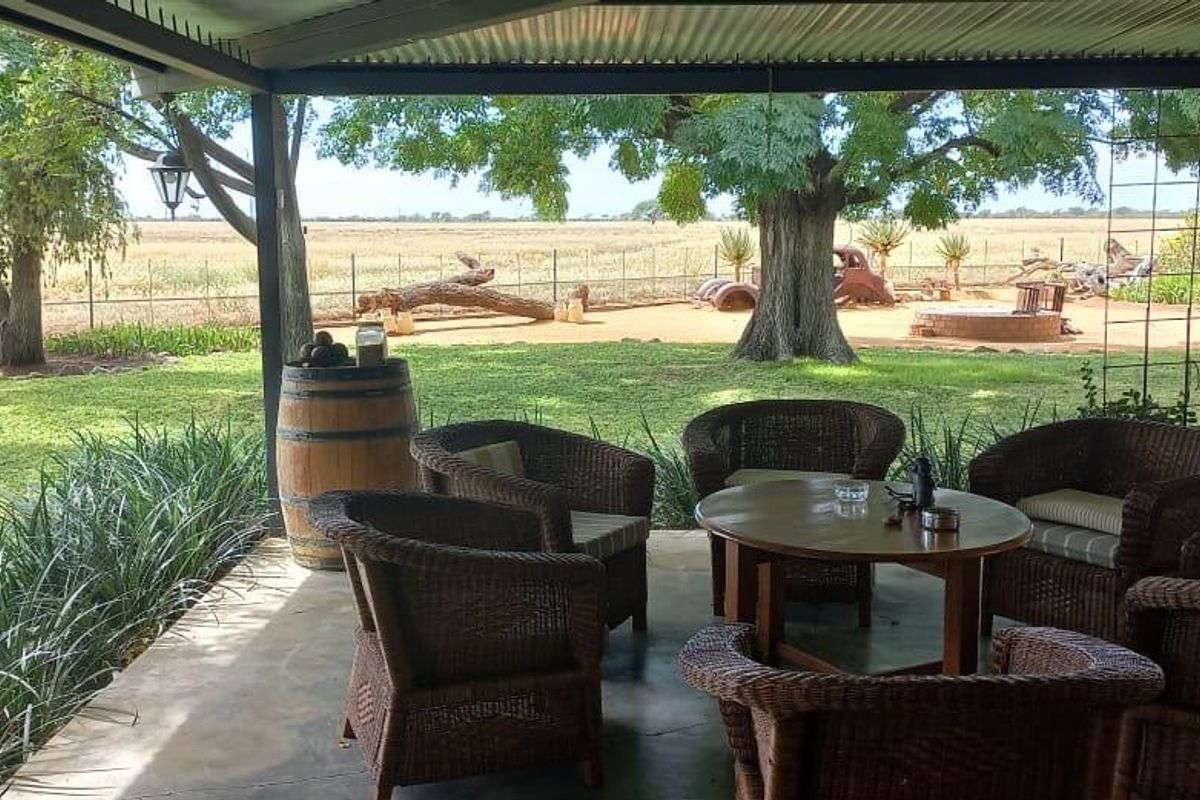 A covered outdoor patio area featuring concrete flooring and metal roofing, overlooking a spacious farm garden and open landscape.
