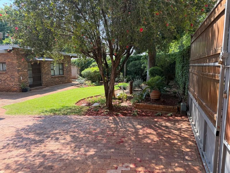A well-maintained garden area featuring brick paving, mature trees, landscaped garden beds, and a brick outbuilding, enclosed by a timber boundary fence.