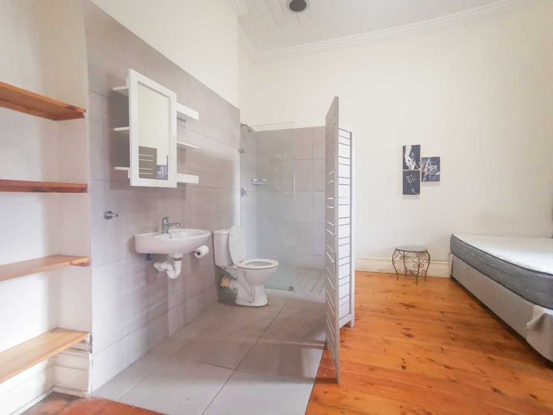 Bedroom featuring an open-plan en-suite bathroom area with a wall-mounted basin, toilet, and glass-enclosed shower, complemented by timber flooring and built-in shelving.