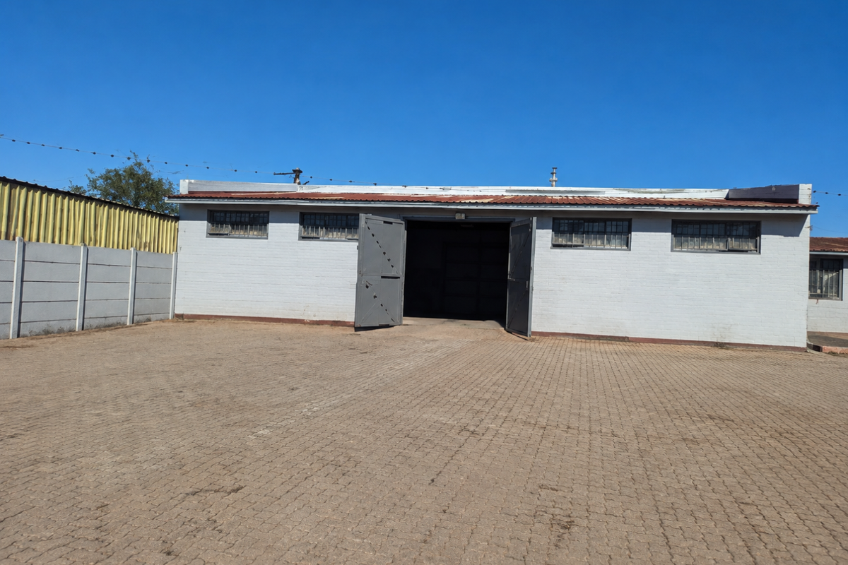 Exterior view of a commercial warehouse building featuring a large paved yard, secure metal entrance gates, and security-barred windows.