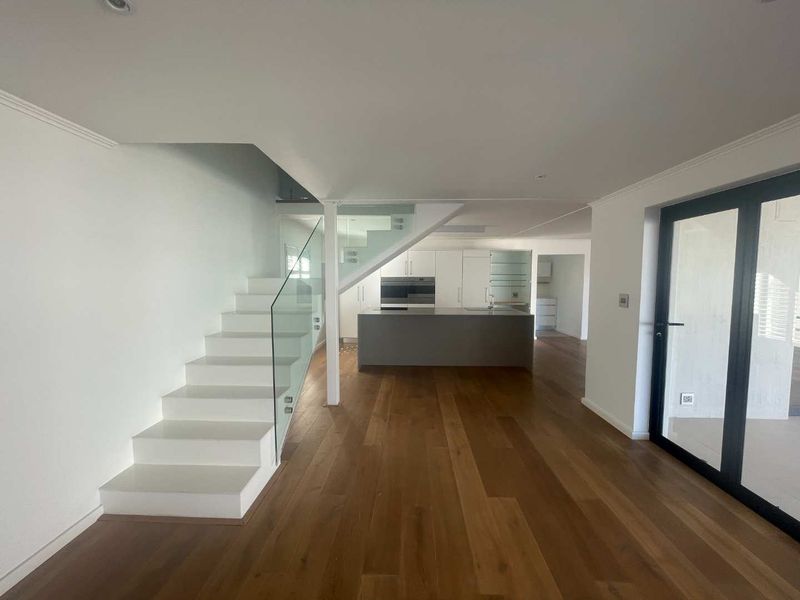 Spacious open-plan living area featuring modern hardwood flooring, a sleek kitchen island with built-in appliances, and a contemporary staircase with glass balustrades.