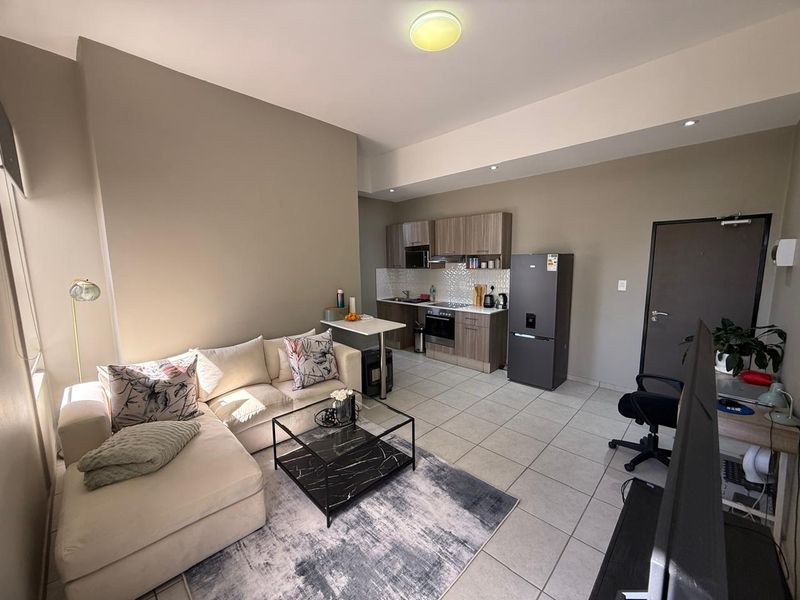 A modern open-plan living area featuring a compact kitchen with built-in cabinetry, integrated appliances, and neutral tiled flooring throughout.